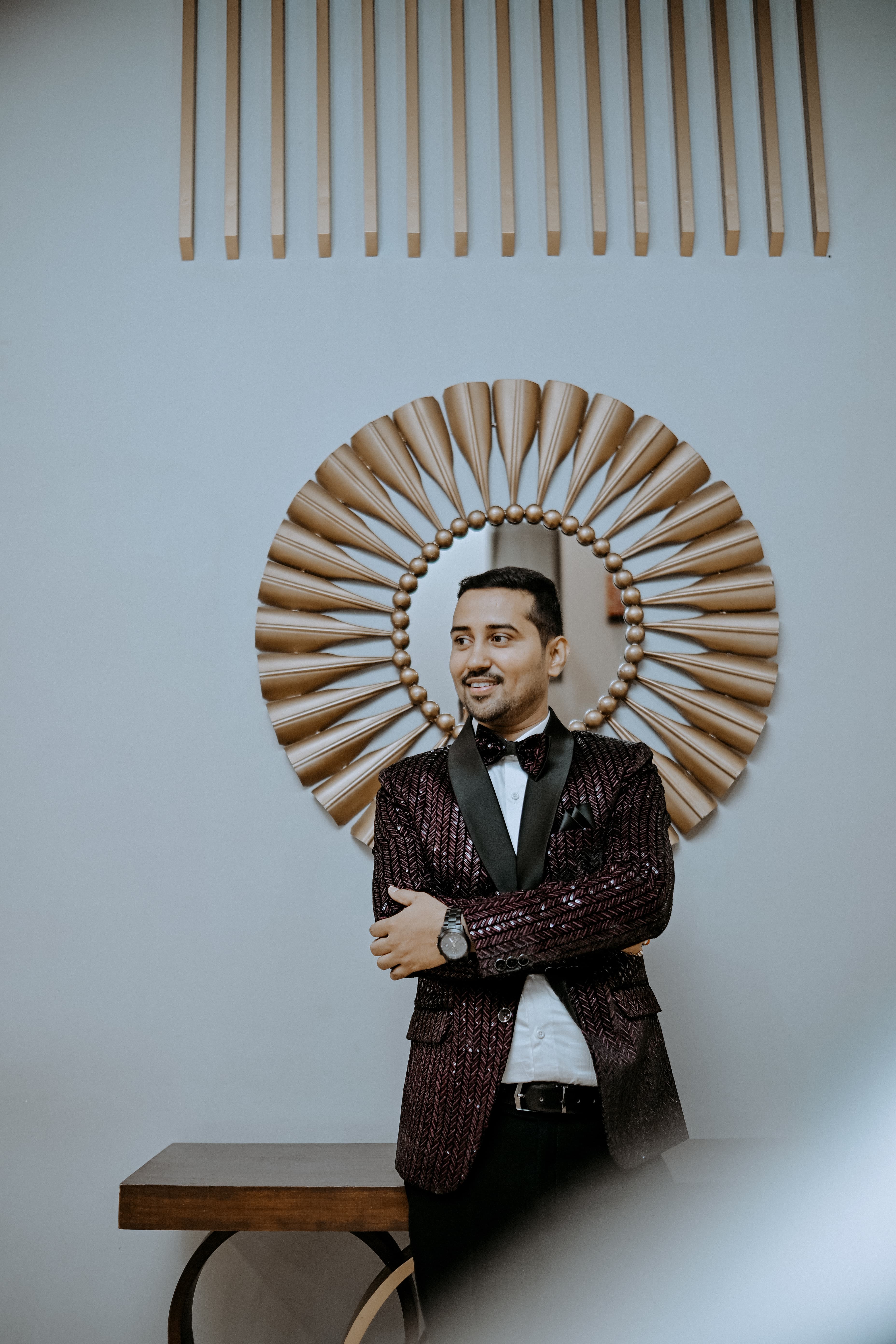 Smiling man in a dark patterned tuxedo posing before a large gold sunburst mirror.